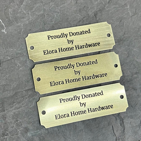 Three brass labels proudly donated by Elora Home Hardware on a gray background
