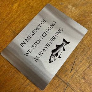 Metal plaque with engraved text and fish illustration on a wooden surface